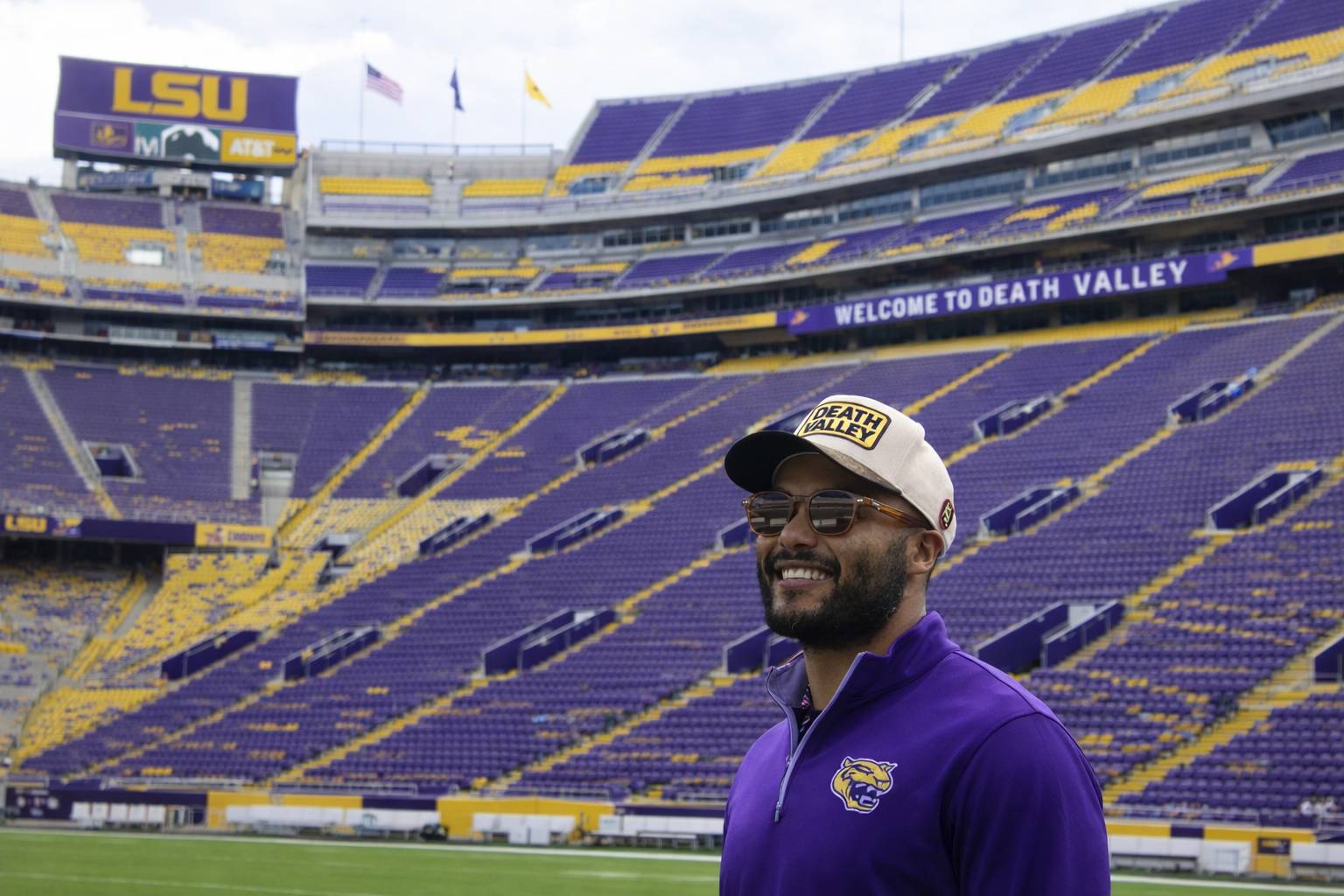Sterling Tiger Valley Man in LSU gear smiles on the field at LSU stadium, with empty purple and yellow stands behind him.